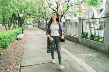 full length young beautiful trendy asian korean girl in long shirt and jeans walking in pathway in park. attractive woman looking aside while carrying backpack. cute female relax outdoor sunny day.