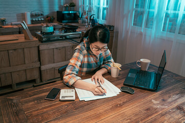 Fototapeta na wymiar college girl student stay up late sitting at table in dark home kitchen at night. young asian korean woman enjoy cup of instant noodles doing homework and preparing for exam with laptop computer.
