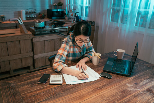 College Girl Student Stay Up Late Sitting At Table In Dark Home Kitchen At Night. Young Asian Korean Woman Enjoy Cup Of Instant Noodles Doing Homework And Preparing For Exam With Laptop Computer.