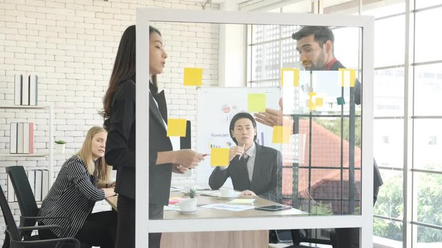 Businesswoman and businessman brainstorming using sticking notes on glassboard discussing ideas with team colleagues. 