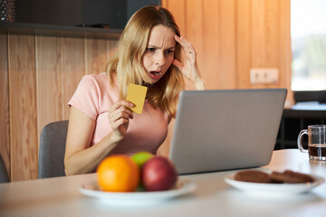 Shocked woman holding credit card and using notebook at home