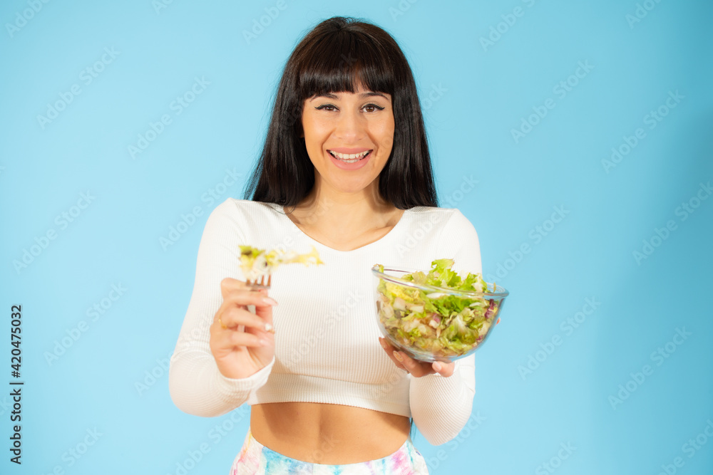 Wall mural portrait of a happy playful woman eating fresh salad from a bowl isolated over blue background