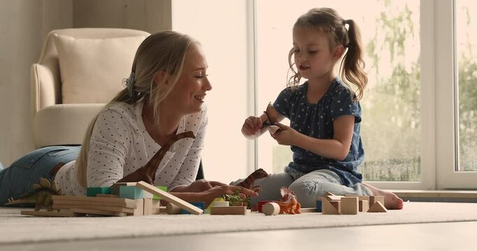 Young woman lying on carpet on warm floor in modern living room communicates play dinosaur toys set with little cute 4s daughter. Family leisure at home, fun and hobby, babysitting and daycare concept