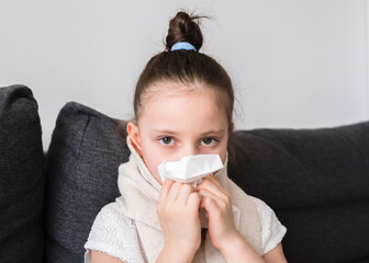 Runny nose, a little girl is being treated at home. Toddler girl sits on the couch and blows her nose into a napkin. The child is cold and sick and has a runny nose and snot. Allergy, flu