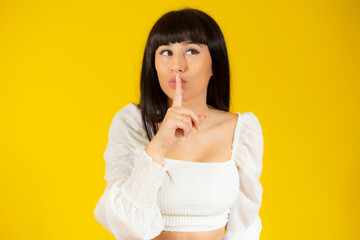 Portrait of beautiful and young brunette woman standing over isolated yellow background asking to be quiet with finger on lips. Silence and secret concept.