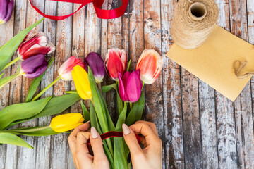 Top view of hands tying rope on colourful tulips bouquet on wooden table.  Florist workplace background. Decorator, diy, craftsmanship, spring gift concept.