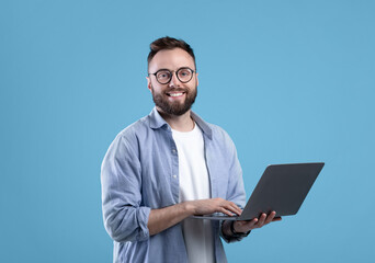 Cheerful millennial guy in glasses using laptop computer for online work or communication on blue...