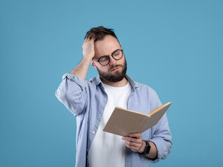 Young Caucasian student having difficulty understanding textbook, studying for exam on blue studio...