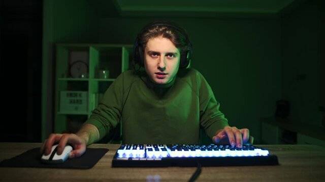 The happy gamer plays video games on the computer at night in a room with a green light and emotionally rejoices in the victory with raised hands and a smile on his face.