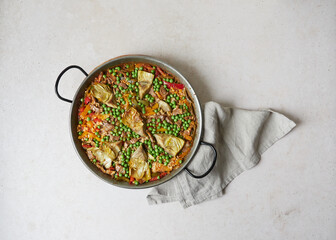 top view of paella cooked in paella pan (traditional Spanish utensil). on gray surface with kitchen towel under the handle.  Vegetable ingredients, peas and artichokes.