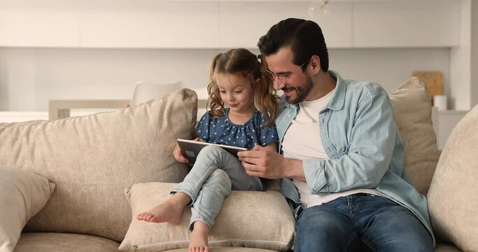 Young father small 4s daughter resting on comfy couch with tablet. Family spend time on internet, small girl sit on soft cushions holding electronic device having fun use app, online amusement concept