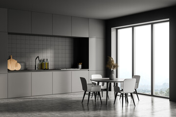 Dark grey modern kitchen interior, dining area with panoramic windows. Furnished by table and chairs for eating. Concrete floor.
