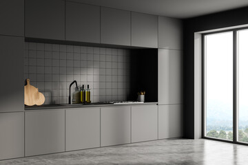 Dark grey modern kitchen interior with kitchenware. Concrete floor and panoramic window with countryside and hills view. Black facades and ceramic tiles on wall