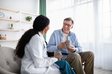 Happy disabled elderly patient and young doctor drinking tea together during medical visit at home
