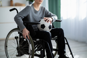 Cropped view of handicapped teen boy with soccer ball sitting in wheelchair, feeling upset over his injury at home