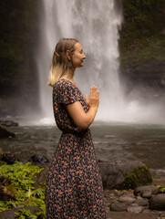 Young Caucasian woman spending time near waterfall. Meditation and relaxation. Hands in namaste mudra. Closed eyes. Travel lifestyle. Woman wearing dress. Nung Nung waterfall in Bali