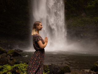 Young Caucasian woman spending time near waterfall. Meditation and relaxation. Hands in namaste...