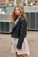 Young stylish woman in pink dress, black leather jacket and boots walking at urban location. Female model with long wavy blonde hair outdoor fashion look.