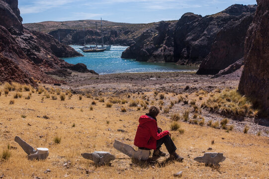 A woman sitting on whale bones looks at the sea and the moored sailboats