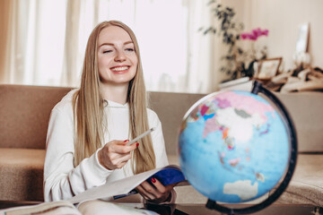 Caucasian girl student smiles, holds a notebook in his hands and studies geography on a globe while...