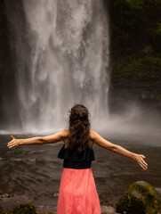 Traveler woman wearing pink dress at waterfall. Excited woman raising arms in front of waterfall. Travel lifestyle. View from back. Copy space. Nung Nung waterfall in Bali