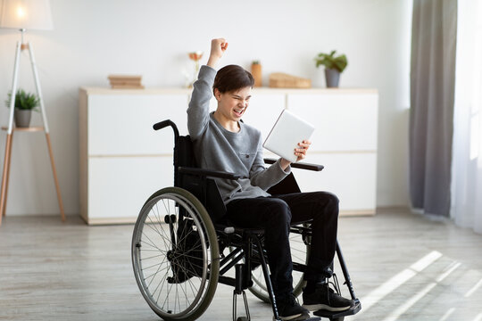 Excited Teen Boy In Wheelchair Holding Tablet Computer, Celebrating His Achievement Or Success, Winning Lottery At Home