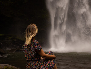 Young Caucasian woman sitting on the rock and enjoying waterfall landscape. Energy of water. Travel lifestyle. View from back. Nung Nung waterfall, Bali