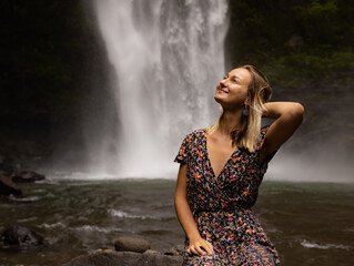 Young Caucasian woman sitting on the rock and enjoying waterfall landscape. Woman portrait. Energy of water. Travel lifestyle. Nung Nung waterfall, Bali