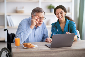 Young nurse showing senior disabled man how to use laptop computer at retirement home