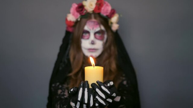 woman in the image of death Santa Muerte or sugar Mexican skull with a burning candle in her hands on a gray background. focus on candle halloween makeup