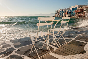 Two empty chairs on the waterfront in the island of Mykonos, Greece. In the background the splash of the waves and the characteristic Greek houses on the coast.