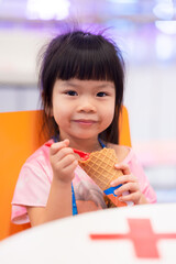 Child girl enjoy eating sweet ice cream cone in summer. Sweet smile kids looking at camera. Tables in front of her were labeled forbidden to sit close to each other, spaced out due to virus outbreak.