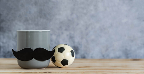Objects on the table of Happy Fathers day holiday background concept.Black mustache on cup with football on blurred grey backdrop.Sign for the season for dad.mock up, border,banner, copy space.