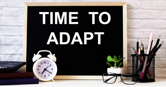 The words TIME TO ADAPT is written on the chalkboard next to the white alarm clock, glasses, potted plant, and pencils in a stand.