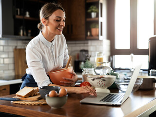 Young woman cooking in the kitchen. Beautiful woman following recipe on laptop and preparing delicious food.