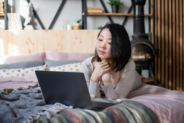 Young Korean woman in home clothes of pastel colors in the bedroom on the bed uses a laptop and relaxes in the interior of the loft and boho. Self isolation. Message and dating  in social net.