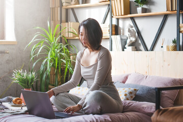 Young Korean woman in home clothes of pastel colors in the bedroom on the bed uses a laptop and...