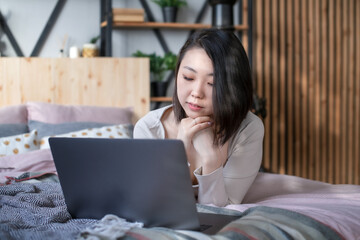 Fototapeta na wymiar Young Korean woman in home clothes of pastel colors in the bedroom on the bed uses a laptop and relaxes in the interior of the loft and boho. Self isolation. Message and dating in social net.