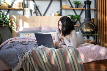 Young Korean woman in home clothes of pastel colors in the bedroom on the bed uses a laptop and...