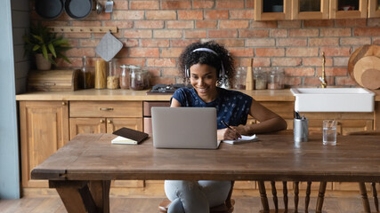Millennial afro american woman in headphones sit by laptop engaged in distant learning foreign...
