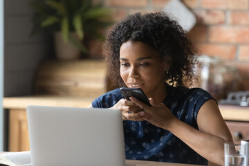 Millennial black female engaged in study work online record thoughts using smartphone app activate digital assistant. Young mixed race woman sit by laptop pc contact customer by cell via voice dialing
