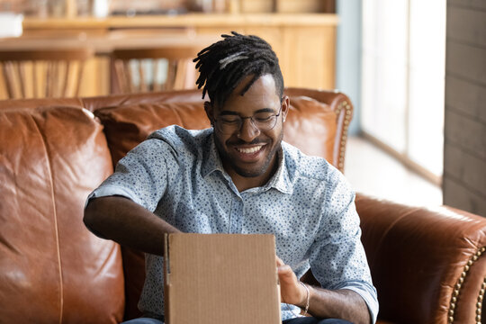Nice thing. Excited black male web shop customer unpack small cardboard box with purchased goods inside. Happy smiling young african man hipster shopper receive delivery by mail enjoy unboxing parcel