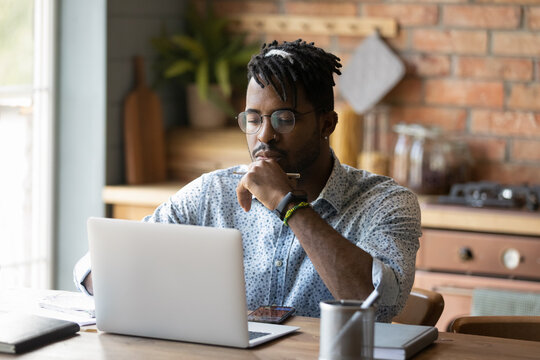 Freelancer At A Job. Young African Man Hipster Think By Laptop At Home Office Prepare Report Electronic Document Use Open Source Web App. Focused Black Male Student Read Web Search Result On Pc Screen