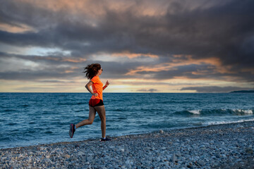 A slender woman in a bright orange T-shirt runs along beach during bad weather.