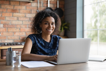 Portrait of smiling biracial female self employed freelancer at work using laptop pc doing research project. Positive motivated young black lady remote student look at camera distracted from learning - obrazy, fototapety, plakaty