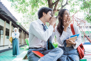 a boy whispered to a girl in high school uniform when sitting in the school garden with angry female friend stand at the background