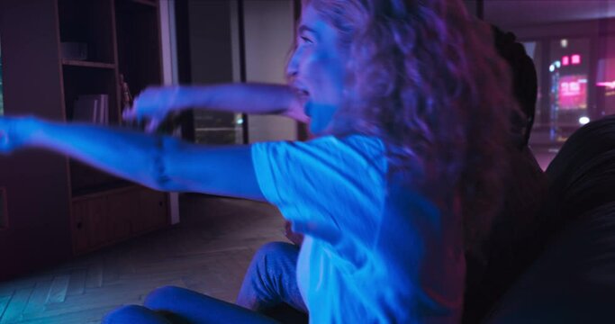 Young man and woman are playing racing video game on TV. They are sitting on the sofa in the living room with red and blue neon lighting. Girl wins and celebrates victory.