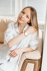 A beautiful blonde girl in a white oversized shirt and blue jeans sits in a bright sunny room on a beige sofa