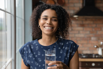 Headshot portrait of smiling young african woman holding glass of fresh clear purified potable water offer to drink. Happy healthy black female look at camera propose you to enjoy clean sparkling aqua