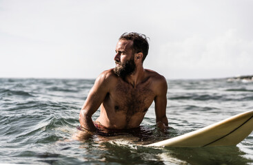 Hipster surfer sitting on his surfboard into the ocean water and waiting for a big wave - Fit bearded man training with surfboard to sea - Adventure and freedom concept doing water extreme sports